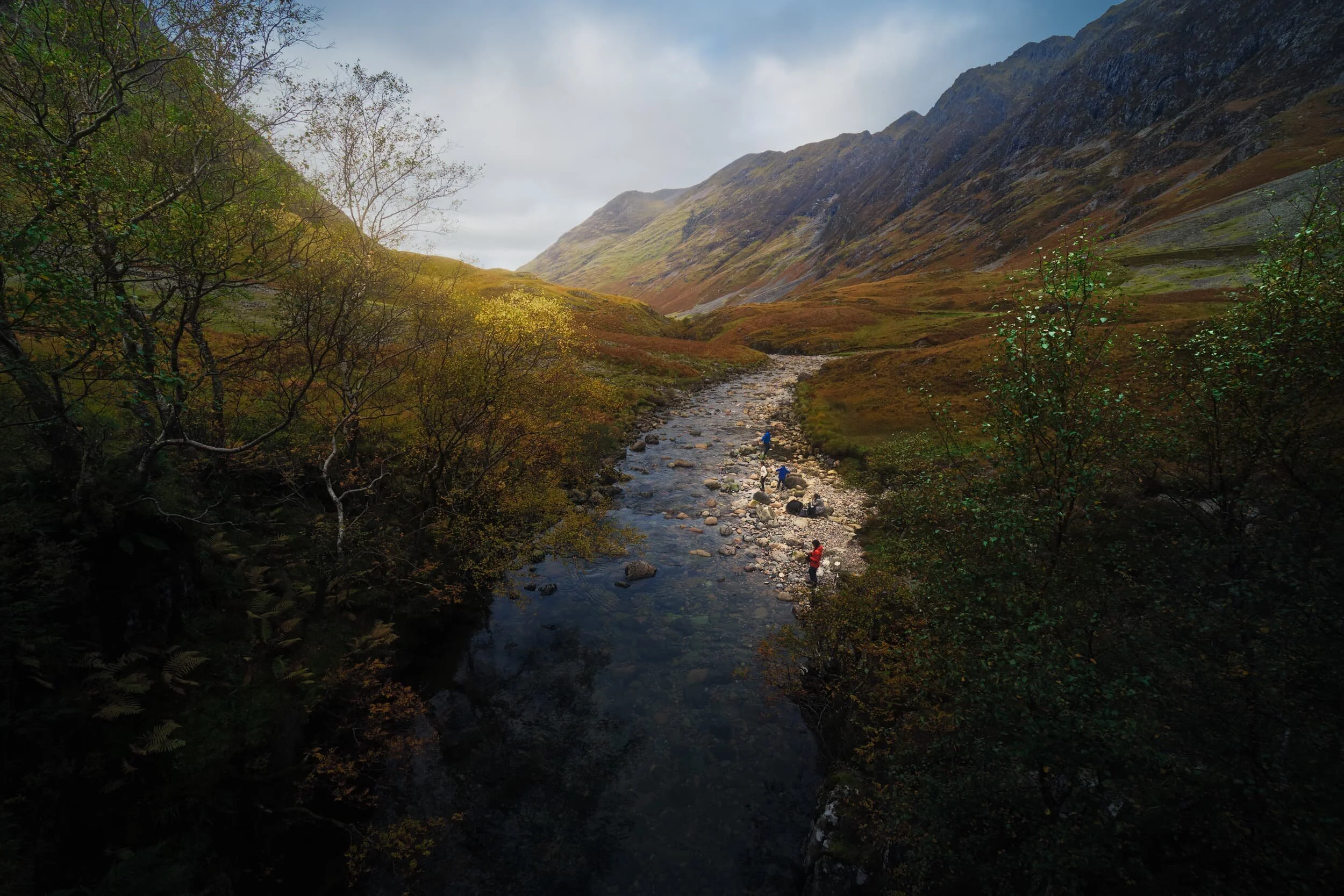 A family play by the calm waters of the River Coe. The giant 10 km ridge in the distance is Aonach Eagach , which rises to 967 m/3,175 ft and borders the northern side of the valley.