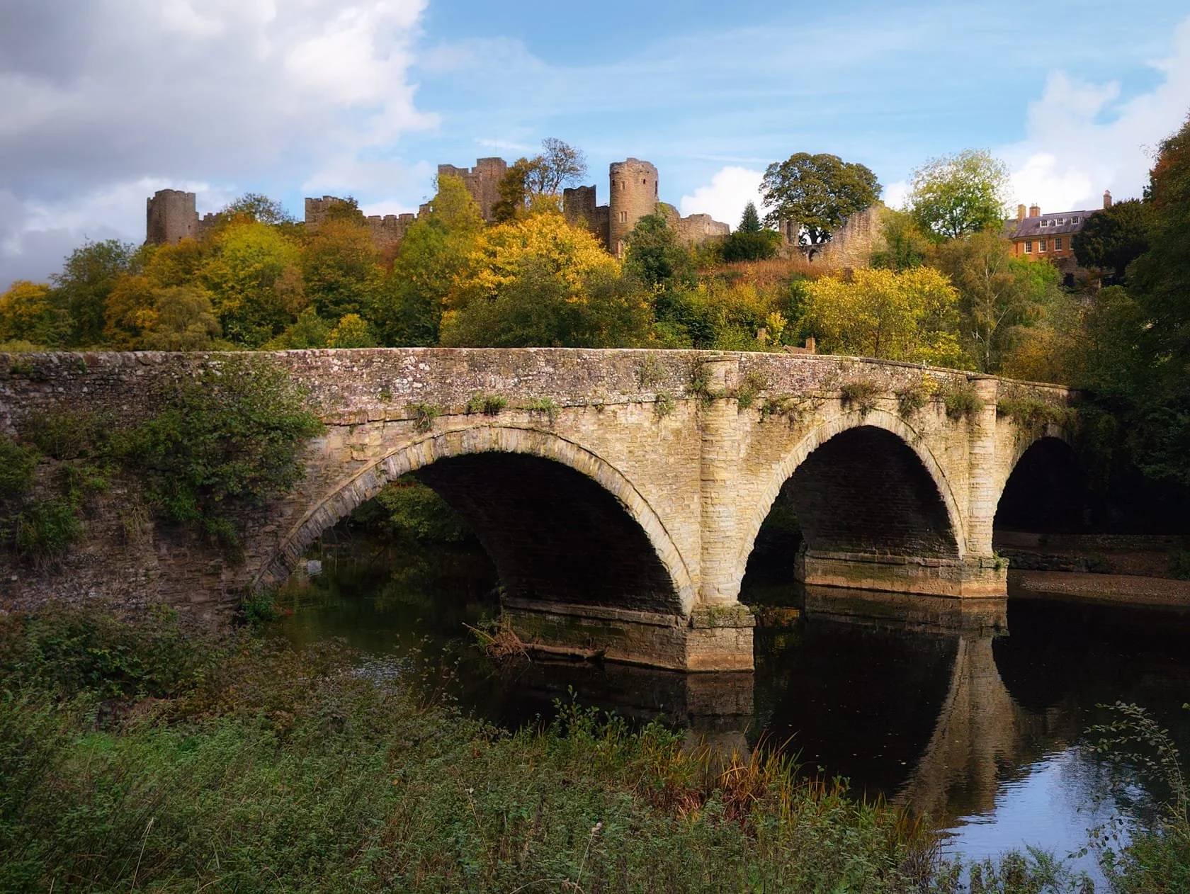 The low sun breaks through the clouds, highlighting Dinham Bridge with the ruins of Ludlow Castle above, adorned in autumnal foliage.