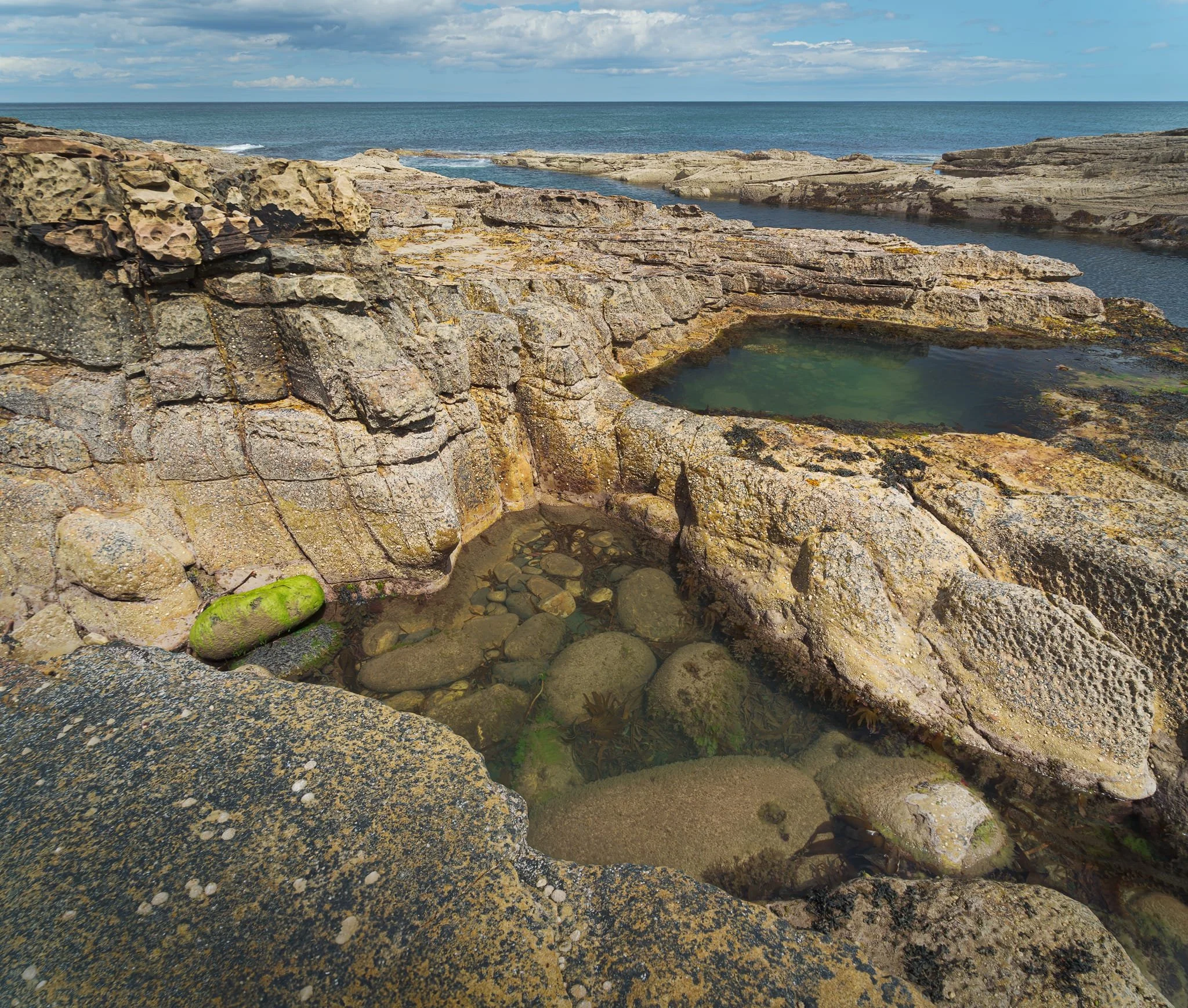 This shot was made from three landscape images at 24mm, stacked top to bottom. Small, circular depressions have been drilled into the sandstone by countless swirls of sand-and-pebble slurry at low tide. The lacy, pitted “bee-nest” pattern on the block tops comes from salt-crystal growth and repeated wet-dry cycles; tiny pockets where salt expands and flakes the rock. Vertical fractures and bedding planes guided where water could exploit weaknesses, prising blocks apart and focusing erosion at those joints and corners.