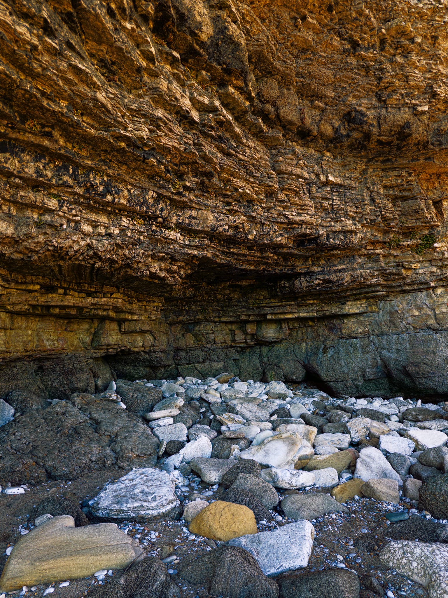 The stunning, layered cliff here is seen in cross-section. Each horizontal band shows a different layer of sediment that was deposited in an ancient, shallow sea. The thinner, finely layered lower beds are followed by thicker, more solid layers above, indicating that the conditions in the water changed over time. Near the base, there’s a prominent overhang, which is a wave-cut notch. Over time, this overhang will likely fall off, and that’s how the boulder beach below was created. The lighter, white stones are fresher pieces of limestone that have recently fallen, while the darker, smoother ones have been shaped and polished by the waves over a longer period.